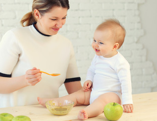 9 month old charming baby sits on the table and mom feeds him with a spoon of applesauce. Blonde hair, blue eyes.