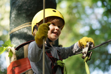 Climber child on training. Cute little boy in climbing safety equipment in a tree house or in a rope park climbs the rope. Safe Climbing extreme sport with helmet - obrazy, fototapety, plakaty