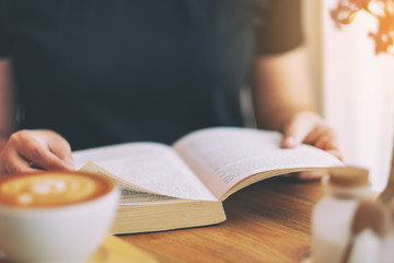 Closeup image of a woman holding and reading a vintage novel book with coffee cup on wooden table