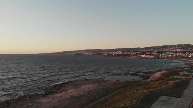 Aerial drone view of mediterranean sea with waves on sunset with city and mountains in background. Seashore with pedestrian path.