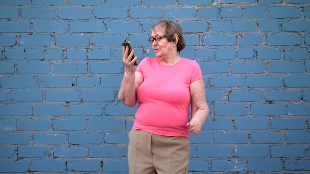 elderly woman in pink T-shirt and glasses dances with new smartphone model and enjoys beautiful quality sound in headphones