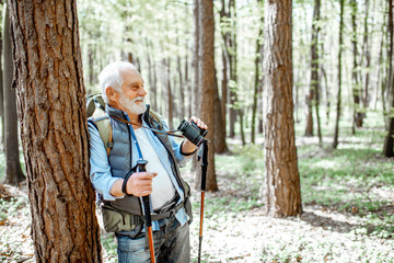 Fototapeta premium Portrait of a senior man with binoculars and backpack resting near the tree while traveling in the forest