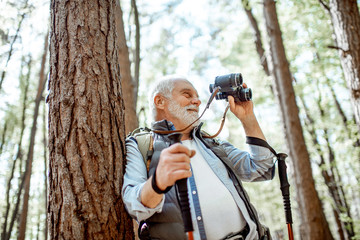 Portrait of a senior man with binoculars and backpack resting near the tree while traveling in the...