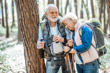 Senior couple resting near the tree while traveling with backpacks and trekking sticks in the forest. Concept of an active lifestyle on retirement