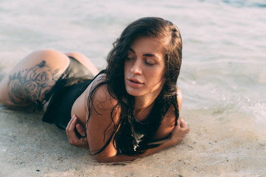 Portrait of sexy brunette girl in bikini and knitted sweater lies on the sandy beach, tattoo on her buttocks.