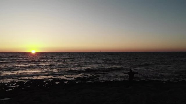 Black dark silhouette of person standing on a beach with arms wide open looking towards sunset over ocean with small boat on horizon. Aerial drone view
