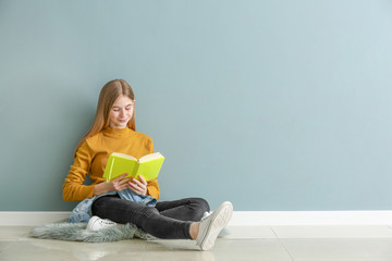 Cute teenage girl with book sitting on floor near color wall