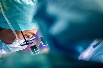 Surgeon performing surgery in hospital operating room. Surgeon in mask wearing loupes during...