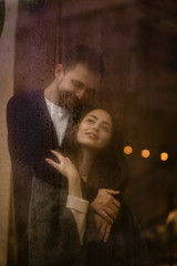 Romantic loving guy hugs his happy girlfriend standing behind a wet window with lights.
