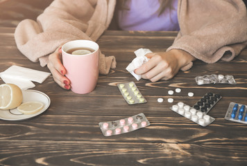 Fototapeta na wymiar ill girl with a flu is sitting at the wooden table with pills and tea. Runny nose and cold. Weak immunity. Winter illness. Lemon and paper tissues.