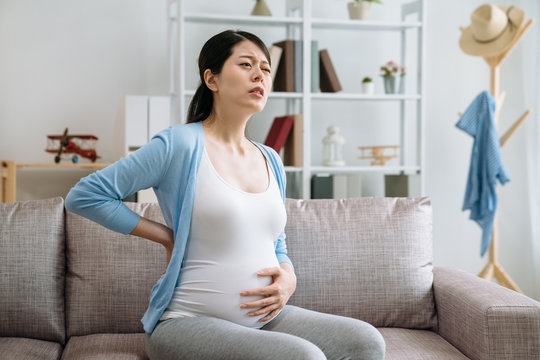 pregnancy health people and expectation concept. young asian pregnant woman in couch touching her back and suffering from backache at home. frowning mother with baby in belly sitting on sofa.