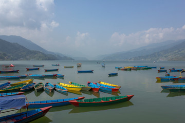 Fototapeta na wymiar many bright multicolored empty wooden boats on the Phewa lake on the background of a green mountain valley in the haze