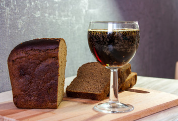 A glass of kvass, a loaf of black bread cut into pieces on a cutting board on a table against the background of a highlighted gray wall. 