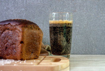 A loaf of black bread on a wooden cutting board and a beige towel and large sea salt on the table with a glass of fresh kvass on a gray background. 