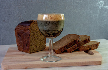 A glass of kvass, a loaf of black bread cut into pieces on a cutting board on a table against a gray background. 