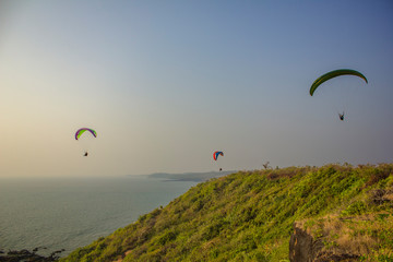 three paragliders on colorful parachutes fly over the sea and the green hill against a clean blue sky