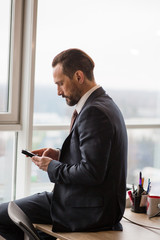 A well dressed businessman is browsing on his mobile phone at the modern office, behind is a window, he is focused and serious.