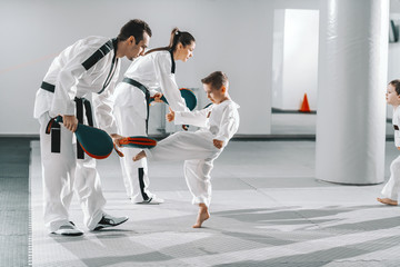 Small group of kids in doboks practicing with their trainers taekwondo moves while kicking in kick...