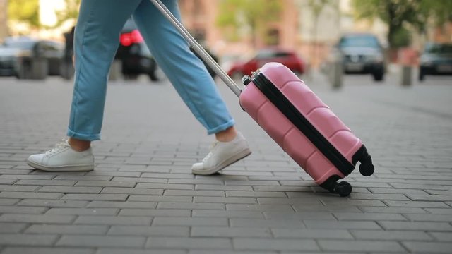 Unrecognizable tourist woman rolls a suitcase goes along sidewalk in a city