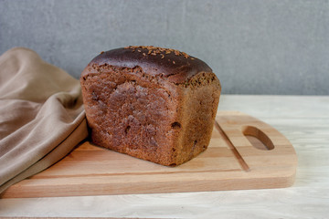 A loaf of black bread on a wooden cutting board and a beige towel on the table on a gray background. 