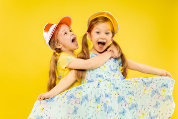 Beautiful emotional little girls isolated on yellow background. Half-lenght portrait of two sisters full of happieness wearing a dresses and caps. Concept of summer, holidays, emotions, childhood.
