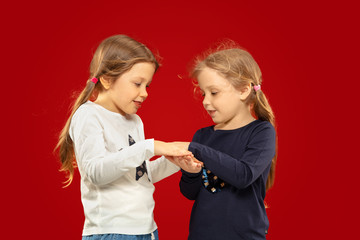 Beautiful emotional little girl isolated on red studio background. Half-lenght portrait of happy sisters or friends standing and smiling. Concept of facial expression, human emotions, childhood.