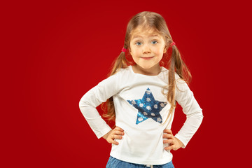 Beautiful emotional little girl isolated on red studio background. Half-lenght portrait of happy child showing a gesture and standing. Concept of facial expression, human emotions, childhood.