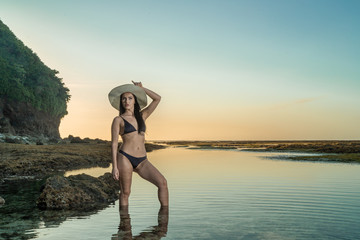Summer vacation happiness carefree joyful woman standing in water enjoying tropical beach destination. Holiday bikini girl relaxing from behind holding straw hat on vacation sea water.
