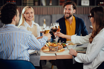 Four happy muliethnic freinds dressed smart casual cheering with alcohol while sitting at restaurant.