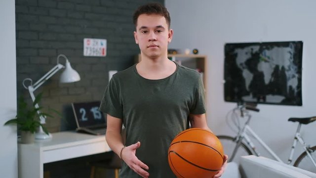 A cute teenager standing with the basketball ball in his room and looking at the camera