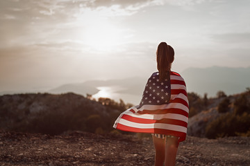 Child teenager girl walks to the top of the mountain with an American USA flag on her shoulders. Sunset time - obrazy, fototapety, plakaty