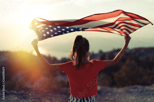 Child girl is waving American flag on top of mountain at sky background. Sunset time