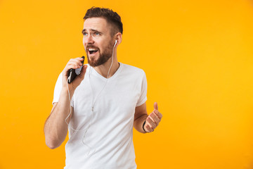 Happy young handsome man posing isolated over yellow wall background using mobile phone listening music with earphones.