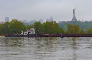 Cargo delivery by river transport. The tug boat towing a barge with sand. The Motherland monument in the background. Spring morning landscape view. Kyiv, Ukraine