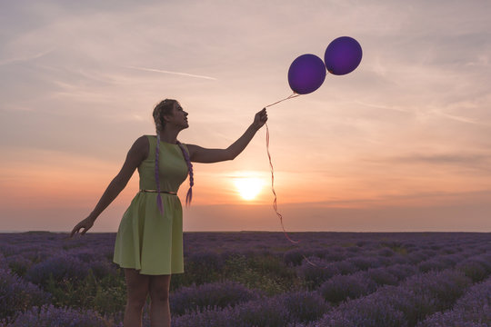 A beautiful model with long braids stands in a lavender field. Holds two balloons in his hands. Sunset in the background. - Powered by Adobe