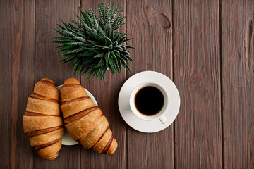 Two croissants on a saucer with black coffee in a white cup on a brown wooden table. Cactus in a pot. Breakfast. Top view. Copy space