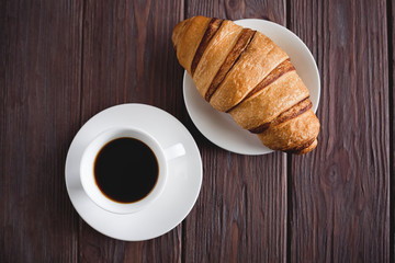 Breakfast. Croissant on a plate and black coffee in a white cup on a brown wooden background. View from above.