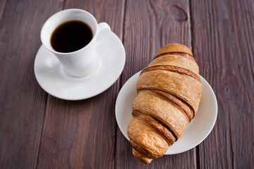 Breakfast. Croissant on a plate and black coffee in a white cup on a brown wooden background.