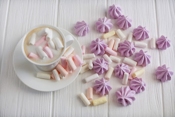 A cup of cappuccino with pastel colored marshmallows on a white table with lilac meringue