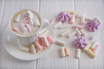 A cup of cappuccino with pastel colored marshmallows on a white table with lilac meringue