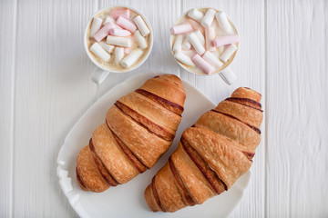 Breakfast for two. Two croissants on a plate, white wooden background with cappuccino and marshmallow, close-up. View from above.