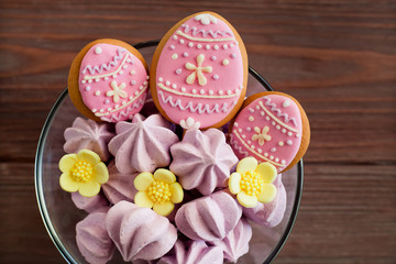 Sweets, meringues and pink gingerbread in the shape of eggs, sugar flowers in a transparent bowl on a brown wooden background
