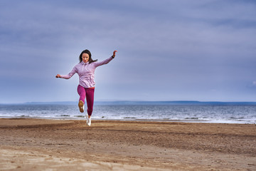 Fototapeta premium Young woman with black hair jumps while jogging. A woman is engaged in gymnastics in the spring morning on the sandy bank of a large river. Cloudy morning.
