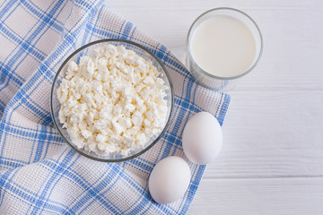 Fresh cottage cheese in a bowl with a glass of milk and a pair of white eggs on a white wooden table, a blue checkered towel. 