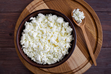 bowl with cottage cheese and a spoon with cottage cheese lies on a wooden board on a brown table, top view