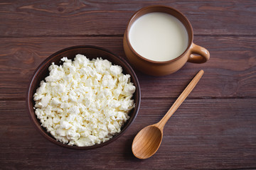 bowl of cottage cheese with a cup of milk with a wooden spoon on a brown table, top view