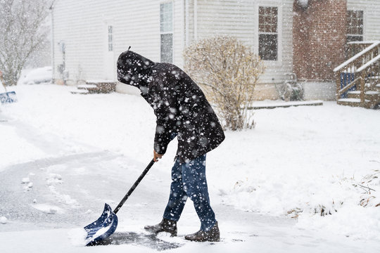 Young Man In Winter Coat Cleaning Shoveling Driveway Street From Snow In Heavy Snowing Snowstorm With Shovel By Residential House And Snowflakes Falling