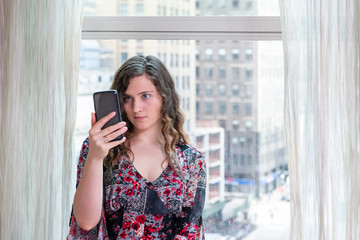 Fototapeta premium Closeup of young happy woman sitting at window in home room looking at phone smiling in NYC taking selfie