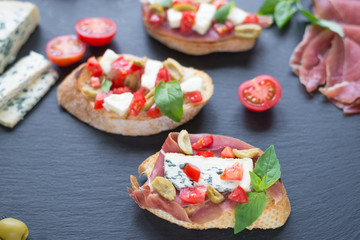 Traditional Italian bruschetta with blue cheese, feta, tomatoes, basil leaves, jamon on black stone background.