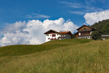 Fototapeta na wymiar A house with flower terraces in a mountainous area against a yellow-green hilly meadow, forest, white clouds, blue sky on a bright sunny day